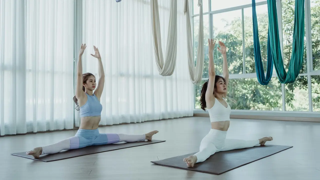 Two women practicing traditional yoga splits pose in a bright studio with aerial yoga hammocks hanging in the background